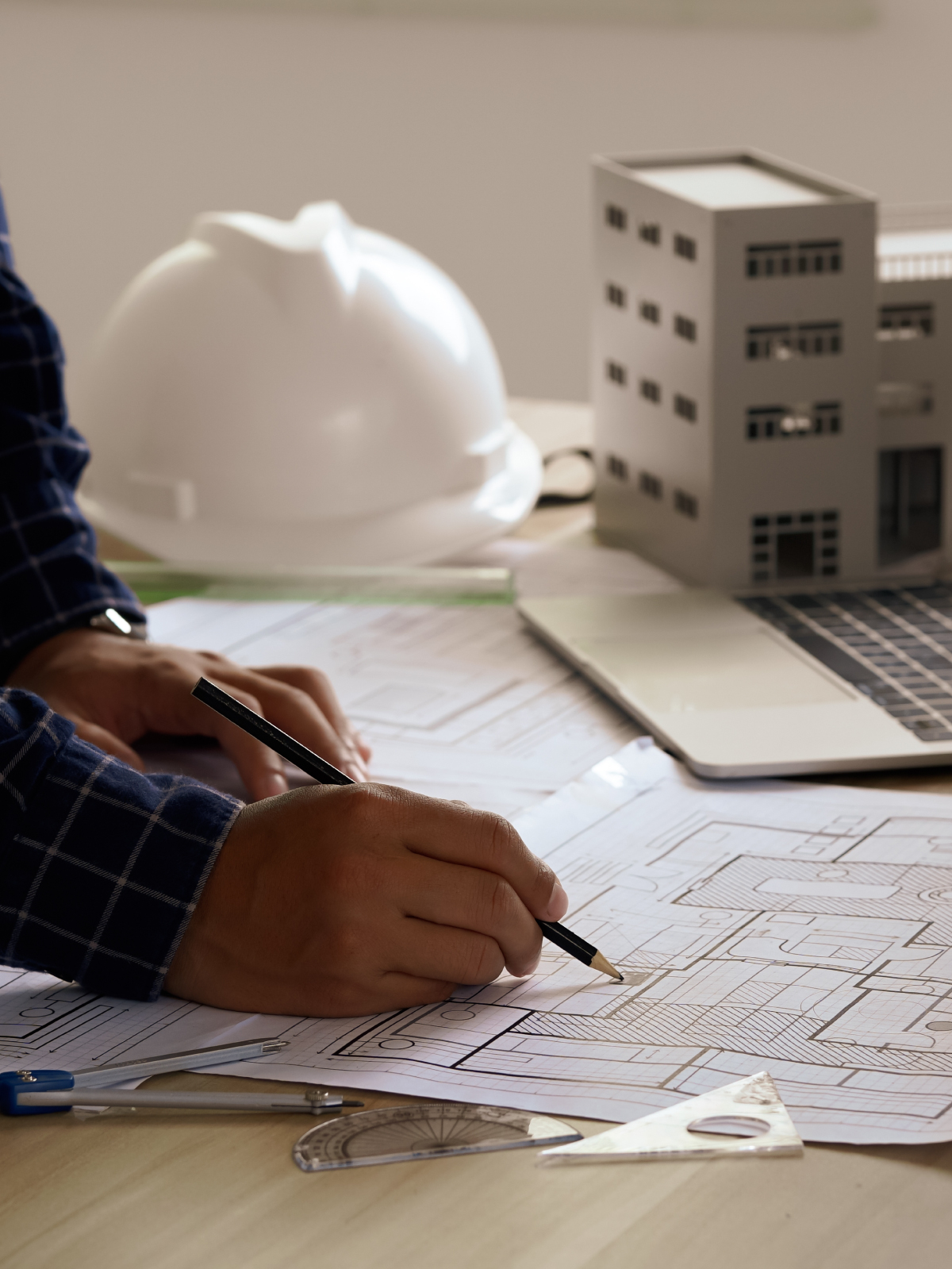 Architect sketching floor plans at a desk with a model building, hard hat, and laptop alongside